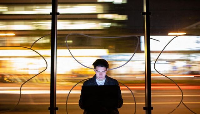 Focused young man working on laptop at night by glass bus stop with neon city light trails