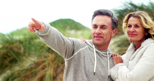 A middle-aged Caucasian man is pointing at something in the distance, with a middle-aged Caucasian woman standing close by, both smiling and enjoying the outdoors. Their casual attire and relaxed demeanor suggest they are sharing a leisurely moment, on a nature walk or sightseeing.