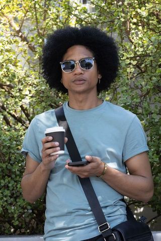 African american man leaning outdoors holding coffee and smartphone while wearing sunglasses