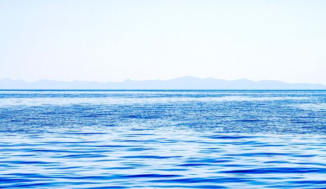 Depicts calm ocean water with distant blue mountains under clear sky. Suitable for use in travel brochures, nature articles, and scenic backgrounds. Conveys tranquility and natural beauty.