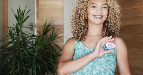 Smiling biracial woman showing vote badge at home, houseplant background