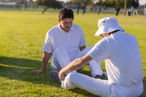 Cricket teammates resting on grass after play, wearing white uniforms and bucket hat