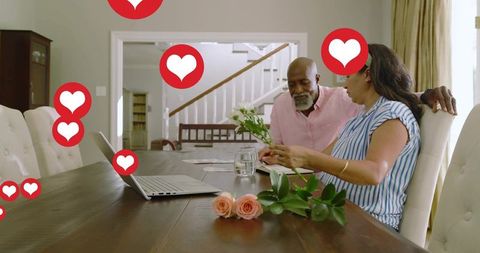 Mature couple arranging roses at dining table while using laptop and sharing tender moment