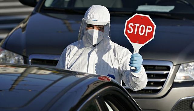 Medical screener in full ppe holding stop sign directing cars at drive-through checkpoint