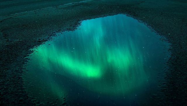 Vibrant aurora borealis reflecting in road puddle on wet asphalt under starry night