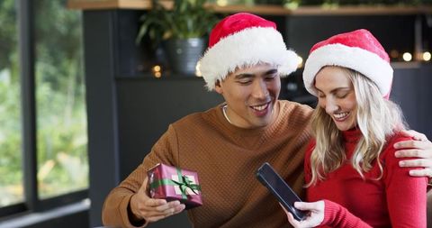 Smiling diverse couple sharing smartphone and wrapped gift in cozy sunlit christmas room