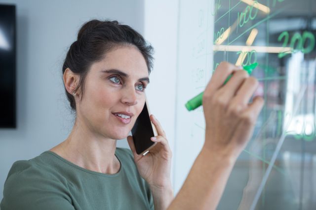 Female executive talking on mobile phone while writing on glass in office