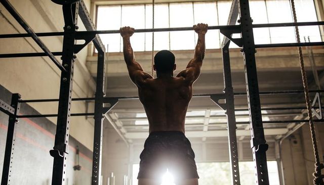 Backlit athlete doing pull-ups in industrial gym warehouse, muscular strength training