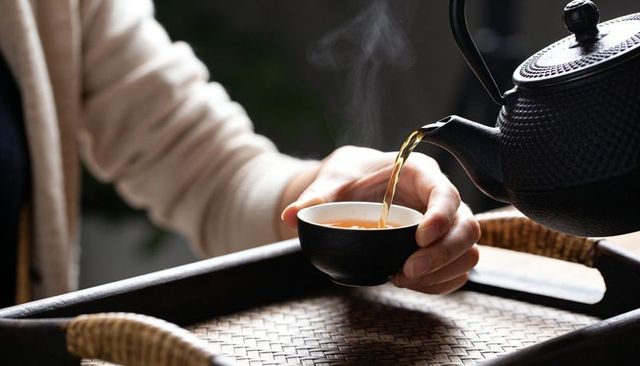Pouring steaming tea into black cup held in hand on rattan tray, cozy warm closeup scene