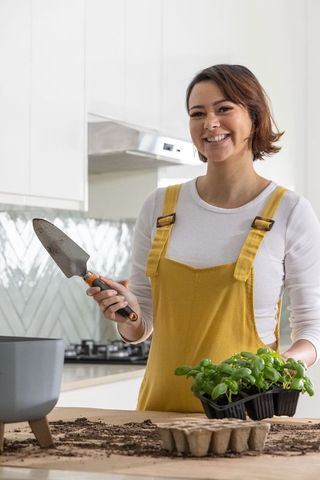 African american woman potting basil in kitchen wearing yellow overalls smiling with herbs