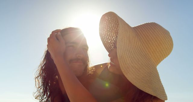 Young Caucasian man and young Biracial woman share a tender moment outdoors. Sunlight flares around them, highlighting a romantic beach setting.