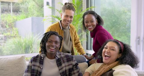 Diverse friends laughing together on sofa in bright living room with garden view
