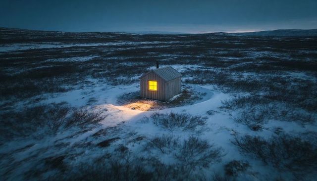 Wooden cabin glowing on snowy moorland at dusk with warm amber window light