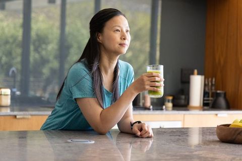 Mature asian woman enjoying green smoothie while leaning on modern kitchen counter