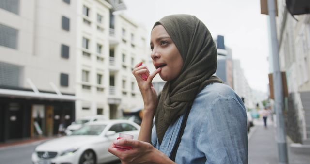 Biracial woman in hijab using lip balm in city street. City living and modern urban lifestyle.