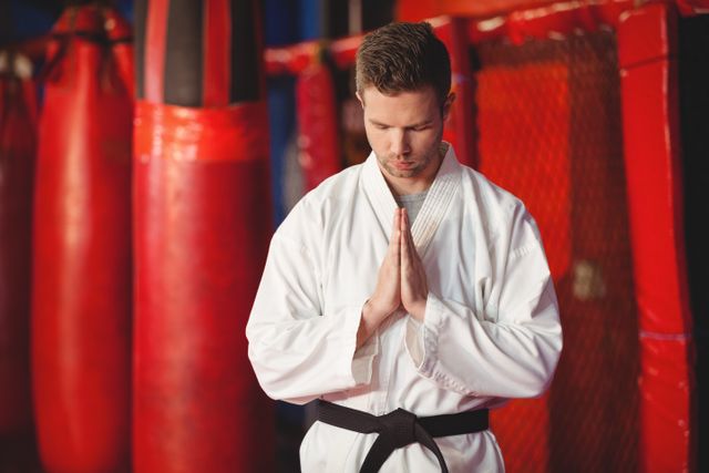 Karate practitioner in white gi and black belt meditating in martial arts studio with red punching bags in background. Ideal for use in articles about martial arts, fitness, meditation, discipline, and self-defense training.