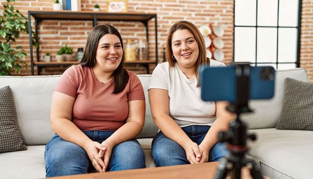 Two friends filming smartphone vlog on sofa smiling casual home lifestyle content creation