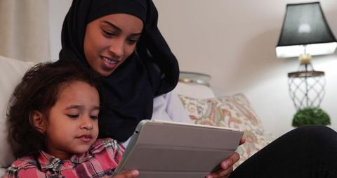 Mother wearing hijab sitting on sofa helping daughter with tablet computer