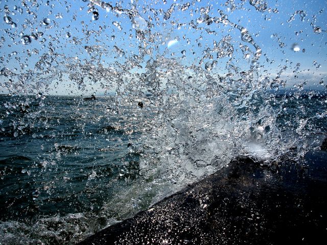 Refreshing and dynamic image of water splashing against a rocky shoreline with calm sea and clear blue sky in background. Captures energy and power of nature, perfect for backgrounds, travel advertisements, presentations about coastal or marine environments, and bringing a touch of freshness and vitality to any visual project.
