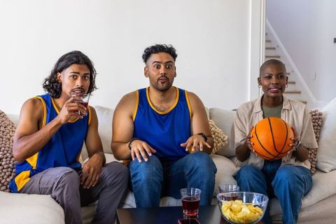African american friends watching basketball game on couch wearing team jerseys holding basketball