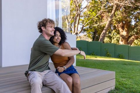 Multiracial couple sitting on backyard deck embracing, smiling and enjoying sunny garden