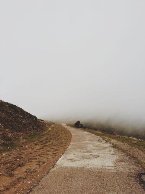 A winding desert road disappears into a foggy horizon, creating a serene and tranquil atmosphere. This image is ideal for use in travel blogs, websites, and promotional materials to evoke feelings of adventure, solitude, and mystery. It can also be utilized in environmental campaigns and inspirational content highlighting nature's unpredictable beauty.