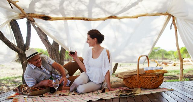 A middle-aged Caucasian couple enjoys a romantic picnic outdoors, with copy space. They are relaxing under a canopy with a wine glass and basket, sharing a moment of leisure in a natural setting.