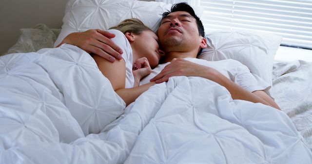 Couple rests closely together under white blankets in a bright, serene bedroom. Suitable for topics related to relationships, relaxation, sleep health, bedroom decor, or domestic lifestyle.