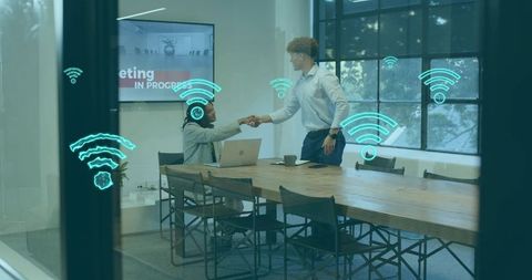 Shaking hands in modern conference room with wireless connectivity icons and laptop