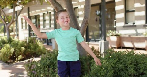 Smiling schoolgirl raising arms enjoying sunlit school courtyard with greenery