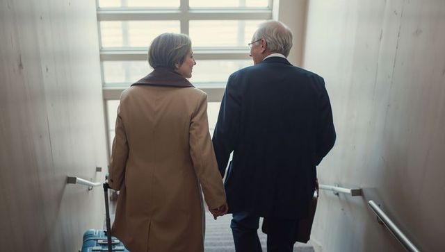Senior couple holding hands walking down interior stairs with rolling suitcase and briefcase