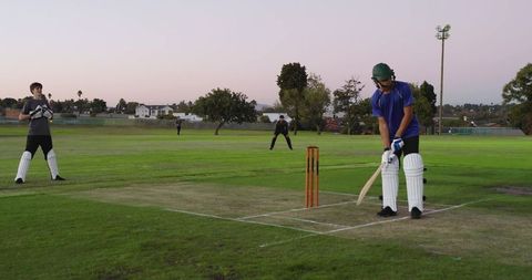 Batsman preparing shot on cricket pitch at dusk wearing green helmet and pads near stumps