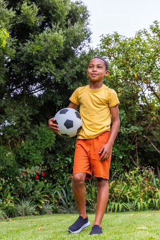 African american boy holding soccer ball in sunny backyard garden smiling and confident
