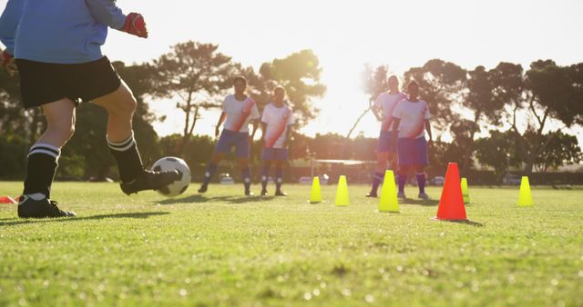 Soccer players wearing uniforms practicing coordinated drills with brightly colored cones on a grassy field as sunlight filters through trees. Ideal for illustrating teamwork, physical training, soccer coaching, outdoor sports activities, and fitness routines.