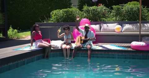 African american friends relaxing on pool deck dipping feet and playing guitar in backyard