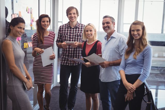 Group of confident business professionals standing together in a modern, creative office. They are smiling and holding documents, suggesting a collaborative and positive work environment. Ideal for use in business-related content, teamwork and collaboration themes, startup promotions, and office culture presentations.