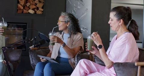 Women recording wellness podcast in cozy lounge holding green smoothies and talking