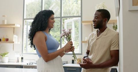 Smiling diverse couple exchanging pink bouquet in bright modern kitchen, intimate moment