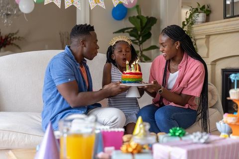African american family celebrating daughter's birthday blowing candles on rainbow layer cake