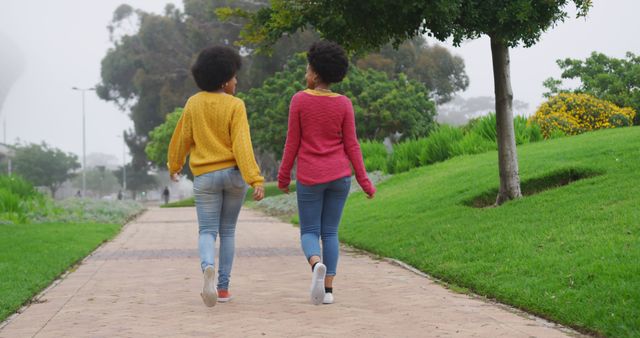 Two women are walking in a park on a foggy day. They are dressed in casual clothing, including sweaters and jeans. The path is surrounded by green grass and trees, offering a serene outdoor environment. This image is suitable for themes related to fitness, nature, friendship, and a healthy lifestyle.