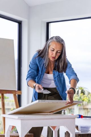 Senior woman smoothing canvas with palette knife in bright home art studio