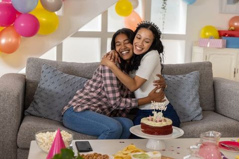 African american mother hugging teen daughter on sofa celebrating birthday with cake