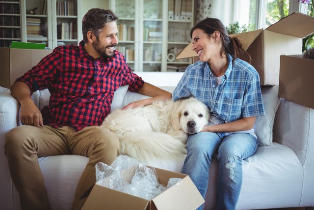 Couple sitting on sofa with their pet dog, surrounded by moving boxes, enjoying a moment of relaxation in their new home. Ideal for use in articles or advertisements about moving, new homes, family life, pet-friendly living, or real estate.