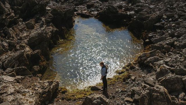 Solo hiker exploring sunlit tide pool amid jagged coastal rocks with backpack and glinting water