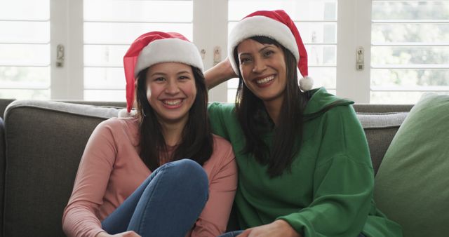 Mother and daughter sitting on a couch in living room, both wearing Santa hats and smiling. Perfect for holiday-themed designs, family-related content, and Christmas promotional materials.