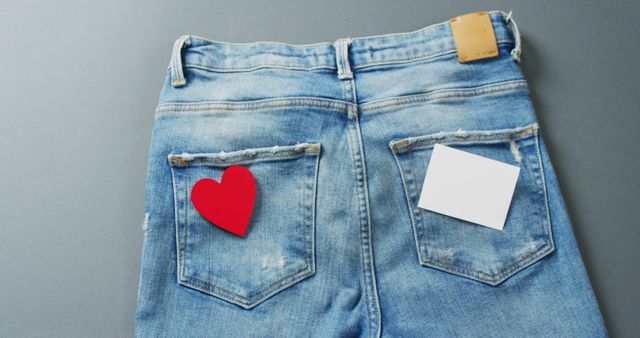 Blue denim jeans are creatively displayed with a red heart and a blank white card placed in the back pockets, symbolizing love and messages. Suitable for use in fashion advertising, clothing catalogs, or Valentine's Day promotions. The image can convey themes like romance, casual style, personal messages, and modern fashion statements.