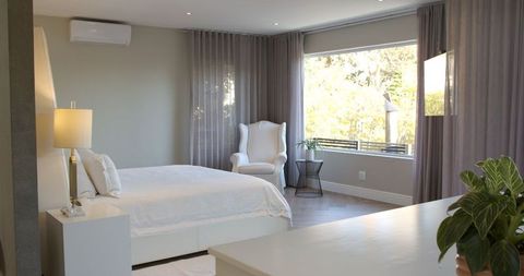 This modern bedroom features a white bed against soft grey walls, exemplifying a minimalist and luxurious design. Natural light floods the space, enhancing comfort and tranquility. The brass table lamp adds an elegant touch, fitting for previews, interior design magazines, or articles emphasizing peaceful and sophisticated lifestyles.