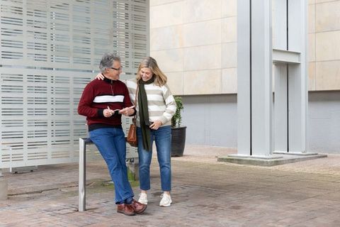 Mature couple sharing smartphone moment while leaning on rail in modern urban plaza