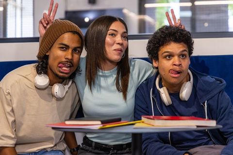 Diverse students laughing and making playful faces in college lounge with notebooks and headphones