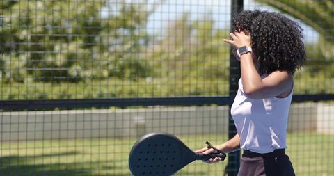 Woman adjusting hair on padel court holding racket while wearing smartwatch